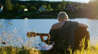 Person spelar akustisk gitarr vid en sjö i lantlig miljö, symbol för folkmusikens tradition, berättande och nära koppling till natur och kulturarv.